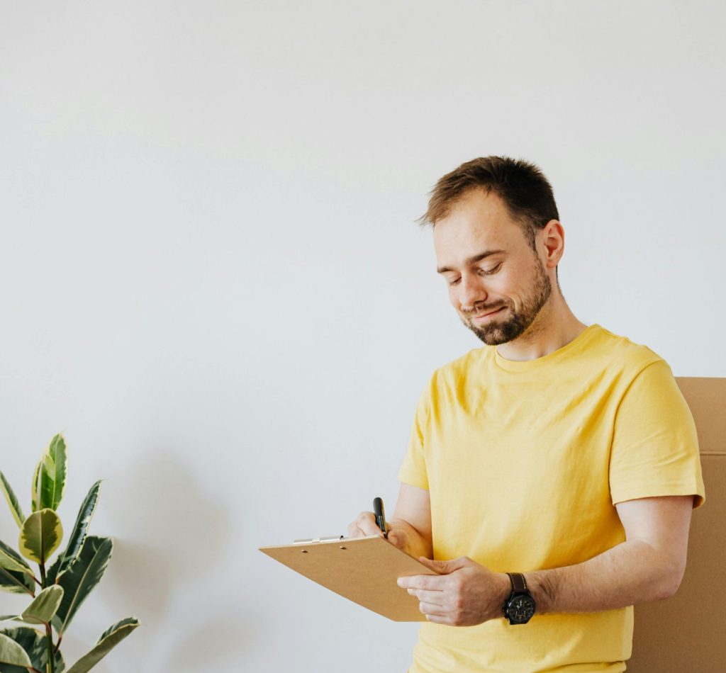 Full body content male in casual wear writing down notes in clipboard while pacing cardboard containers to relocate into new apartment