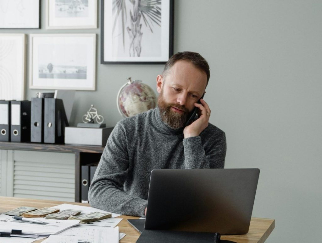 Businessman at home office using laptop and phone, surrounded by office supplies, showcasing a productive remote work setup.