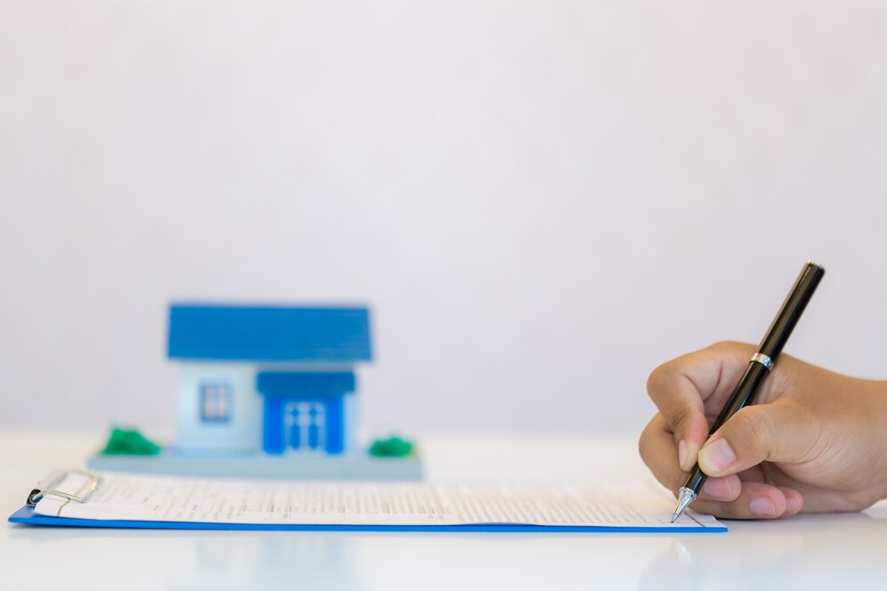 close-up of a hand signing a contract on a table, accompanied by a miniature house model, representing home ownership