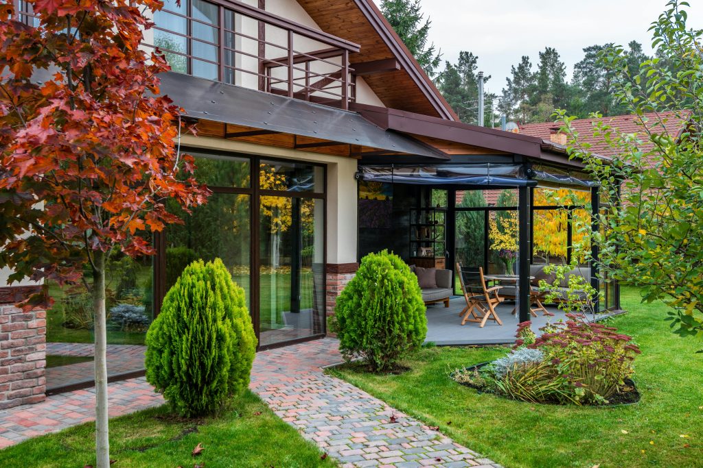 A modern house with large glass doors opens to a lush garden. Red and green foliage, a brick path, and cosy patio furniture create a serene, inviting atmosphere.